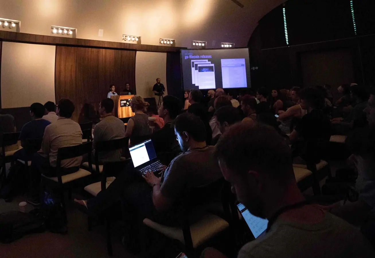 Audience seated in a dark room watching a presentation on a large screen at a tech conference.